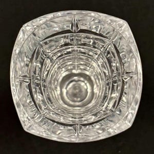 May include: Clear crystal vase with a square top and a circular interior. The vase has a textured, geometric design and is set against a black background. The vase is empty and appears to be made of glass.