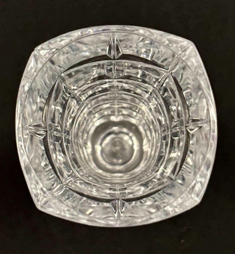 May include: Clear crystal vase with a square top and a circular interior. The vase has a textured, geometric design and is set against a black background. The vase is empty and appears to be made of glass.
