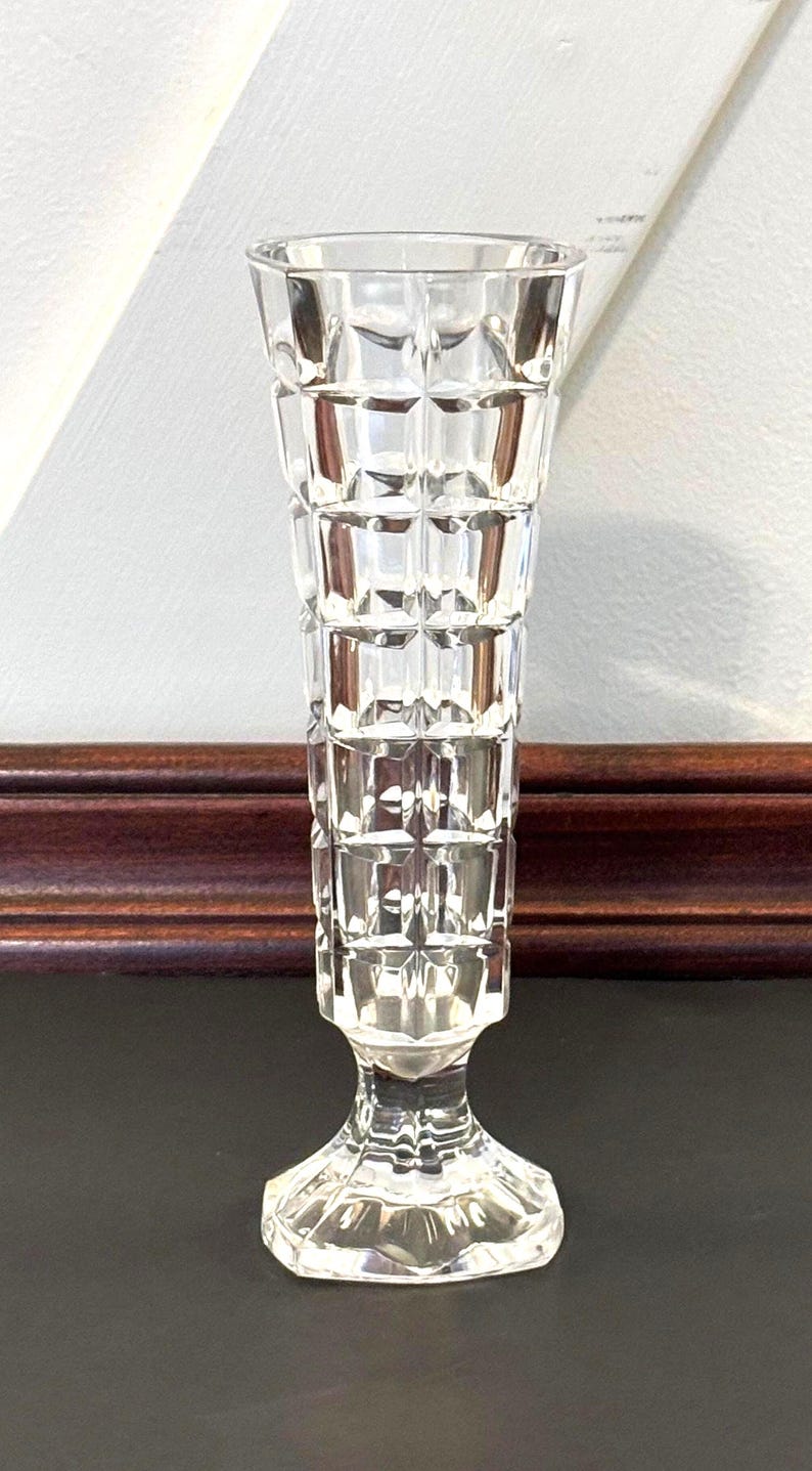 May include: A clear, faceted glass vase with a flared top and a pedestal base. The vase has a geometric, grid-like pattern and is approximately 8 inches tall.