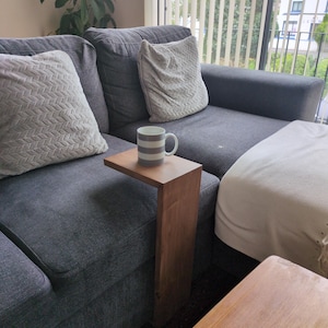 May include: A wooden side table, C-shaped, stands beside a grey sofa. A mug with grey stripes rests on the table. Two decorative cushions and a white blanket are also visible. The table is approximately 40 cm in height.
