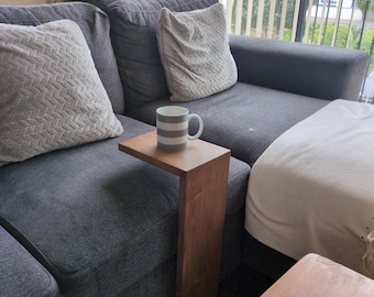 May include: A wooden side table with a C-shaped design is positioned next to a gray sofa. A mug with gray stripes sits on the table. Two decorative pillows and a white blanket are also visible.