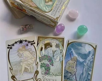 Może przedstawiać: Talia kart tarota "Ethereal Visions" i trzy karty tarota z ilustracjami w stylu Art Nouveau i złotymi detalami. Na zdjęciu widoczne są również różne kamienie szlachetne i przezroczysty kryształ kwarcu.