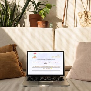 K&ouml;nnte beinhalten: Ein Laptop mit spanischem Text, der auf einer hellen Oberfl&auml;che liegt, mit einem braunen Kissen links und einem rosa Kissen rechts. Der Hintergrund zeigt Topfpflanzen und dekorative Gegenst&auml;nde.