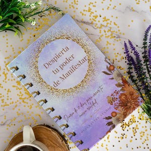 K&ouml;nnte beinhalten: Ein Spiralbuch mit dem Text "Despierta tu poder de Manifestar" auf dem Einband. Das Buch hat ein lila-goldenes Design mit floralen Akzenten. Eine Tasse Kaffee und dekorative Blumen sind im Hintergrund.