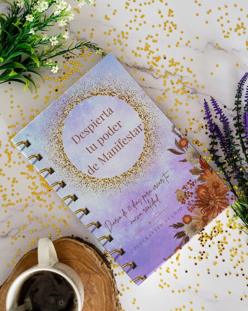 K&ouml;nnte beinhalten: Ein Spiralbuch mit dem Text "Despierta tu poder de Manifestar" auf dem Einband. Das Buch hat ein lila-goldenes Design mit floralen Akzenten. Eine Tasse Kaffee und dekorative Blumen sind im Hintergrund.