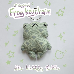 May include: A 3 inch weighted frog keychain made of light green fabric with a dark green fan-shaped pattern. The frog has black embroidered eyes and a small mouth. It is attached to a silver metal keyring. Text reads 3 inch Weighted Frog keychain and the Wobbly Robin.
