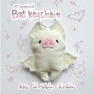 Op de afbeelding: Een 7,6 cm verzwaarde crèmekleurige stoffen vleermuis sleutelhanger met een roze neus, kleine zwarte ogen en twee witte hoektanden. De tekst 3" weighted Bat keychain en the Wobbly Robin zijn zichtbaar op de achtergrond met roze tekeningen.
