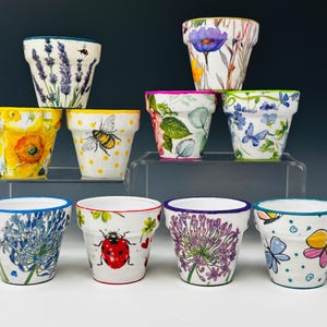 Op de afbeelding: Een verzameling van tien kleine, witte keramische bloempotten. Elke pot is versierd met kleurrijke bloemen- en insectenontwerpen, waaronder lavendel, bijen, zonnebloemen, vlinders en lieveheersbeestjes. De randen zijn in verschillende kleuren geschilderd, zoals blauw, roze en groen.
