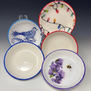 May include: A collection of small, white ceramic bowls with colorful designs. One bowl features a blue bird, another has a string of Christmas lights, and a third showcases purple flowers and a bee. Each bowl has a colored rim.
