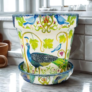 May include: A decorative ceramic flower pot with a matching saucer. The pot features a peacock design with blue, green, and yellow feathers, surrounded by vines, birds, and floral accents. The rim and saucer have a blue border.