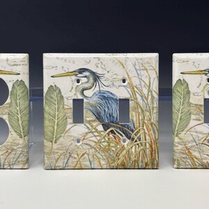 Pode incluir: Coberturas decorativas para interruptores de luz com uma ilustração em aquarela de uma garça azul em um ambiente pantanoso. As coberturas têm um fundo de mapa vintage e estão disponíveis em configurações simples, duplas e de tomada.