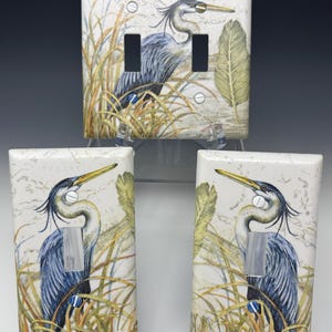 Könnte beinhalten: Dekorative Lichtschalterabdeckungen mit einer detaillierten Illustration eines Blauen Reihers in einer Sumpflandschaft. Der Reiher ist mit blauen und grauen Federn, einem gelben Schnabel dargestellt und von hohem Gras und einem großen Blatt umgeben. Die Abdeckungen sind weiß mit einer strukturierten Oberfläche.