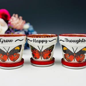 Könnte beinhalten: Eine weiße Keramik-Tasse mit schwarzem Henkel und dem Text "But First, Coffee" in schwarzer, handschriftlicher Schrift. Die Tasse steht auf einer weißen Oberfläche.