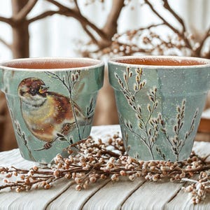 May include: Two decorative flower pots. One pot features a detailed painting of a sparrow perched on a branch, with brown, gold, and white accents. The other pot has a similar design with a floral pattern. Both pots have a teal background.