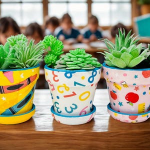 May include: Three decorative flower pots with succulents. The pots are painted with colorful designs: yellow with crayons, white with numbers, and pink with apples and pencils. Each pot sits on a matching saucer.