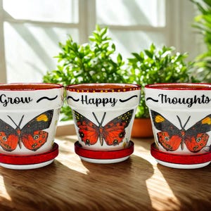 May include: Three white ceramic flower pots with red rims and saucers. Each pot features a butterfly design with orange, black, and yellow wings. The pots are inscribed with the words "Grow", "Happy", and "Thoughts".