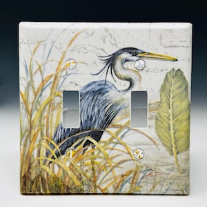 May include: A square double light switch cover plate featuring a detailed illustration of a blue heron standing among tall yellow and green grasses. The background displays a faint vintage map design with text reading Atlantic Ocean.