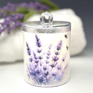 Op de afbeelding: Een cilindrische container met een zilveren deksel en een heldere, bolvormige handgreep. De container is versierd met een aquarelontwerp van lavendelbloemen in paarse en blauwe tinten, met twee kleine bijen. De achtergrond is wit.