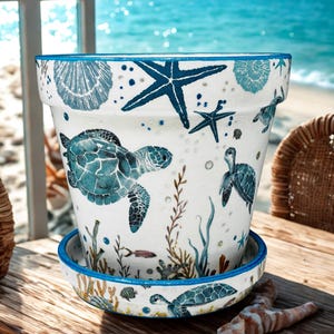 May include: White ceramic plant pot with a matching saucer, decorated with blue sea turtles, starfish, and seashells. The pot has a blue rim and is set against a beach backdrop. A decorative item for coastal or nautical themed decor.