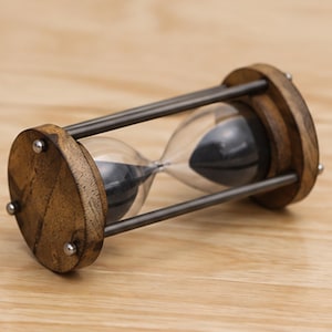May include: An hourglass featuring two circular wooden end caps with a natural grain, connected by four dark metal rods. The central glass bulb contains fine black sand, with the hourglass positioned horizontally on its side to display the structure.