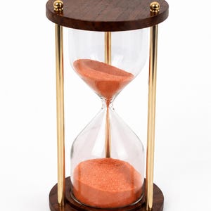 May include: An hourglass featuring a clear glass bulb filled with orange sand, supported by two dark brown circular wooden bases and three vertical gold-toned metal rods. The top wooden base is accented with three small, spherical gold-toned metal finials.