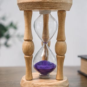 Könnte beinhalten: Eine Sanduhr mit einem hellen Holzrahmen, bestehend aus einem runden Ober- und Unterteil, die durch vier gedrechselte Säulen verbunden sind. Der zentrale Glaskolben enthält leuchtend violetten Sand, der sich größtenteils unten angesammelt hat.