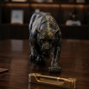 Op de afbeelding: Een zwart panterbeeldje met een gespikkelde gouden afwerking, afgebeeld in een sluipende houding. Het beeldhouwwerk heeft gedetailleerde gezichtscontouren en spierdefinitie, met goudkleurige spikkels verspreid over het donkere oppervlak.