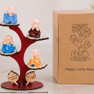May include: A red wooden tree-shaped display stand with four small monk figurines in blue, maroon, and orange robes, measuring 7.5 inches tall and 5 inches wide, accompanied by a brown cardboard box labeled Happy Little Monks with a tree illustration.
