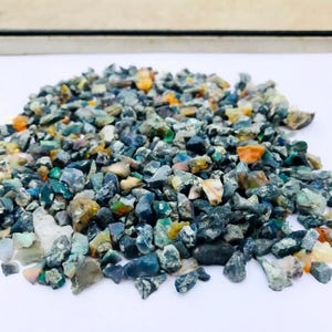 May include: A collection of raw opal gemstones, in various sizes and shapes. The stones exhibit a spectrum of colours, including blues, greens, oranges, and whites. The opals are scattered on a white surface, highlighting their natural, rough texture.
