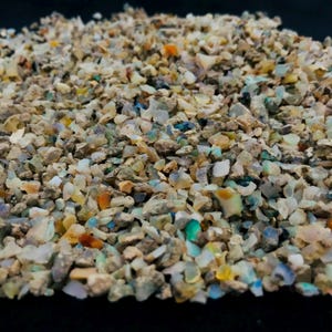May include: A close-up of a collection of small opal chips. The chips are a mix of colours, including white, tan, blue, green, and orange. The texture is rough and granular, with a combination of opaque and translucent pieces.