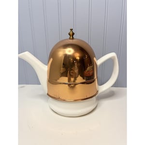 Può includere: Una teiera vintage con base e manico in ceramica bianca, sormontata da un coperchio in rame lucido. Il coperchio in rame ha un piccolo ornamento decorativo. Il beccuccio è bianco e il design è classico e retrò.