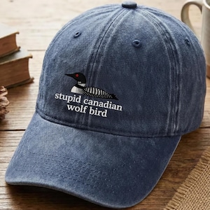 May include: A blue denim baseball cap with a curved brim. The cap features a graphic of a bird with the text "stupid canadian wolf bird" in white. The cap is on a wooden surface.