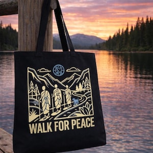 Könnte beinhalten: Schwarze Tragetasche mit einem Naturmotiv. Das Design zeigt drei Figuren, die auf einem Pfad gehen, Berge, Bäume und die Worte "WALK FOR PEACE" in Weiß. Die Tasche hängt an einem Holzpfosten, im Hintergrund ein See und Sonnenuntergang.