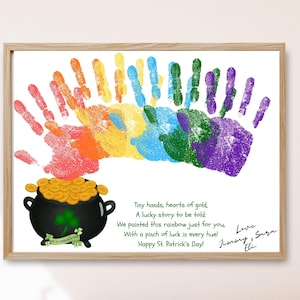 Puede incluir: Impresión enmarcada con un arcoíris de huellas de manos de colores. Debajo, una olla negra llena de monedas de oro y un trébol. El texto incluye "¡Feliz Día de San Patricio!" y firmas.