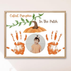 Puede incluir: Impresión enmarcada con el texto "Cutest Pumpkin In The Patch". La impresión presenta una calabaza de acuarela, vides verdes, huellas de manos naranjas y una foto circular de un bebé. El marco es de madera clara.