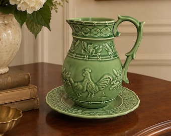 May include: A light green ceramic pitcher and matching saucer set featuring embossed rooster and foliage motifs. The pitcher has a curved handle and a decorative rim pattern, while the saucer displays a detailed border design in matching green.