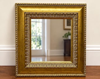 May include: A rectangular wall mirror featuring a wide, ornate gold frame with a beaded outer edge and a detailed, repeating carved pattern along the inner border. The reflective glass surface is clear and smooth, set within the metallic frame.