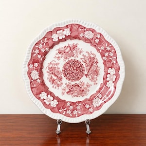 May include: A round ceramic plate with a scalloped edge featuring a red and white transferware pattern. The design includes a central floral medallion surrounded by intricate botanical motifs and geometric lattice borders.