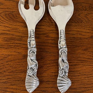 May include: A pair of silver-toned metal salad serving utensils consisting of a three-pronged fork and a rounded spoon. The handles feature a detailed, raised relief design of various seashells, including conch and scallop shells, extending down.