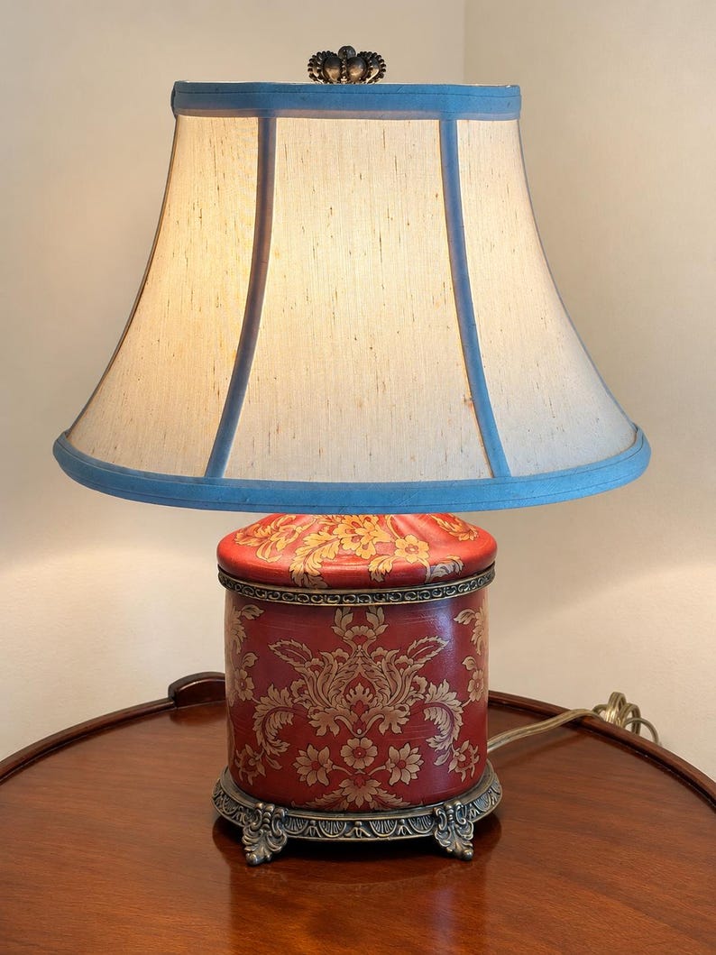 Może przedstawiać: Lampa stołowa z cylindryczną czerwoną podstawą w złoty wz&oacute;r damasceński oraz ozdobną metalową n&oacute;żką. Lampa posiada rozszerzany, kremowy abażur z jasnoniebieskim wykończeniem, zwieńczony dekoracyjnym metalowym elementem na szczycie.
