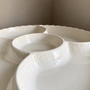 May include: A cream-colored ceramic serving dish featuring a central circular compartment surrounded by three larger, curved sections. The outer edges and the walls of the compartments are decorated with a raised, repeating leaf-like pattern.
