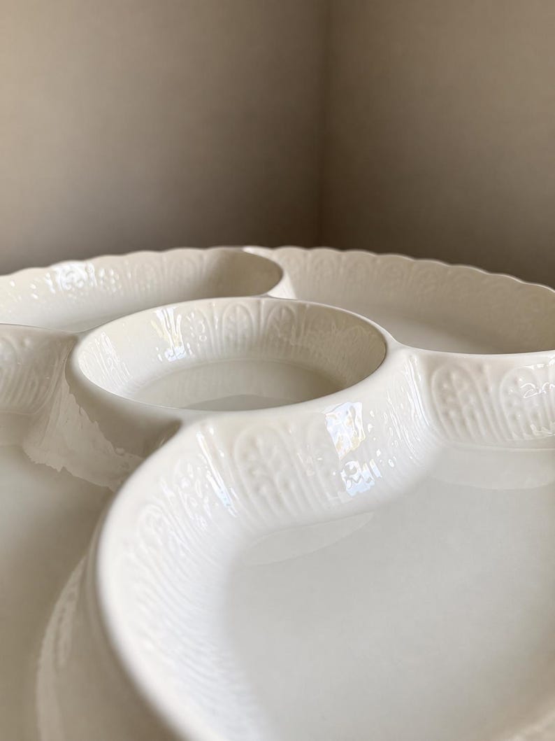May include: A cream-colored ceramic serving dish featuring a central circular compartment surrounded by three larger, curved sections. The outer edges and the walls of the compartments are decorated with a raised, repeating leaf-like pattern.