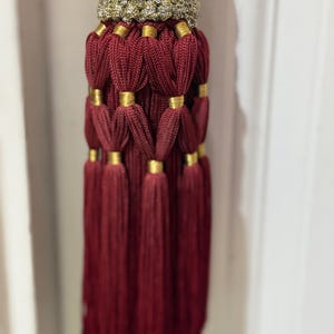 May include: A decorative tassel featuring deep burgundy cord construction with a textured gold metallic band at the top. The design includes multiple layers of looped burgundy cord segments secured by small gold metallic rings, ending in a long fringe.