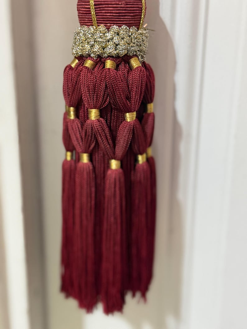 May include: A decorative tassel featuring deep burgundy cord construction with a textured gold metallic band at the top. The design includes multiple layers of looped burgundy cord segments secured by small gold metallic rings, ending in a long fringe.