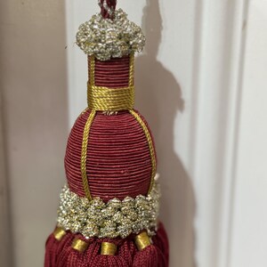 May include: A decorative tassel featuring a deep burgundy cord and skirt, accented with metallic gold thread wrapping and a textured silver and gold braided collar. The top is finished with a silver and burgundy twisted hanging cord.