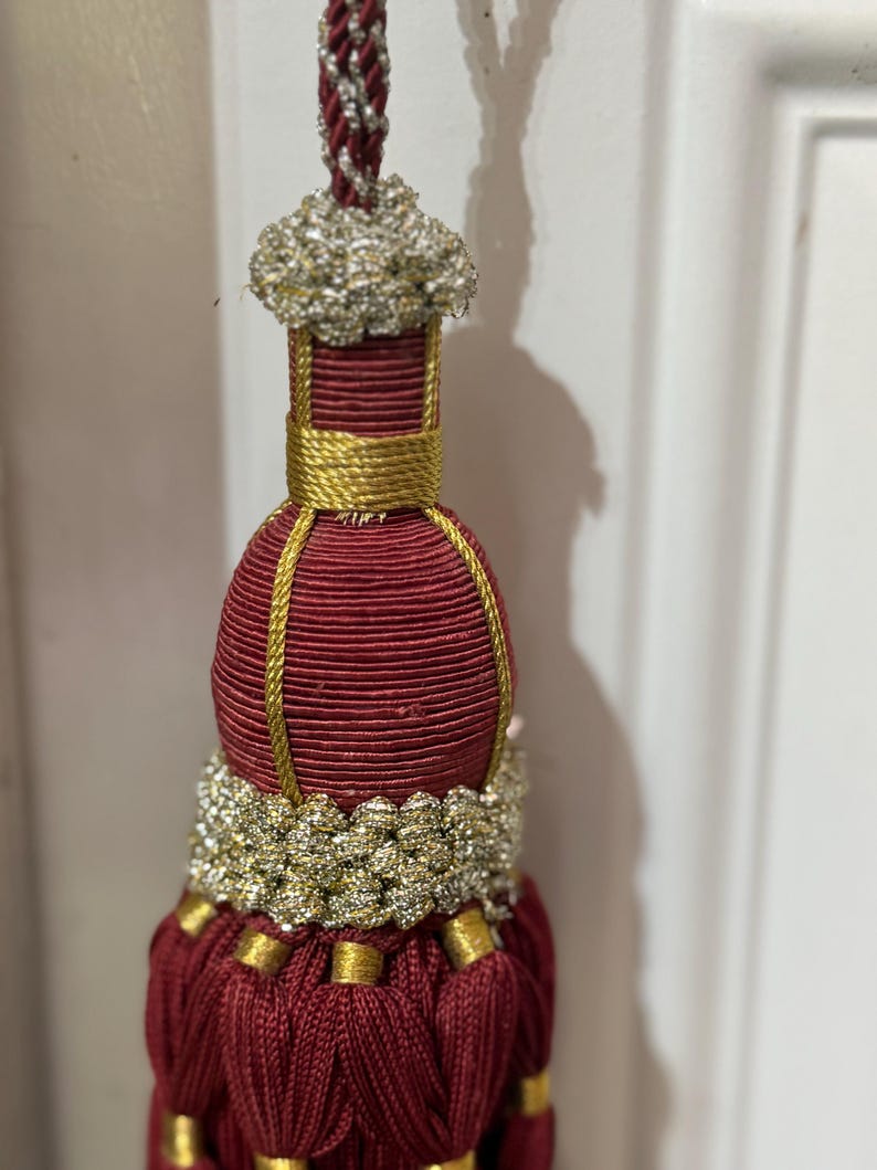 May include: A decorative tassel featuring a deep burgundy cord and skirt, accented with metallic gold thread wrapping and a textured silver and gold braided collar. The top is finished with a silver and burgundy twisted hanging cord.