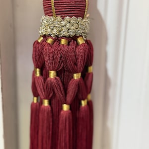 May include: A decorative tassel featuring a deep burgundy corded top section with a gold braided trim. Below this, a textured gold band sits above multiple layers of burgundy cord loops secured by small gold rings, ending in long, straight fringe.