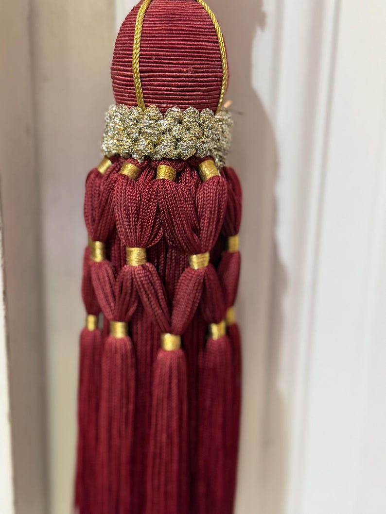 May include: A decorative tassel featuring a deep burgundy corded top section with a gold braided trim. Below this, a textured gold band sits above multiple layers of burgundy cord loops secured by small gold rings, ending in long, straight fringe.