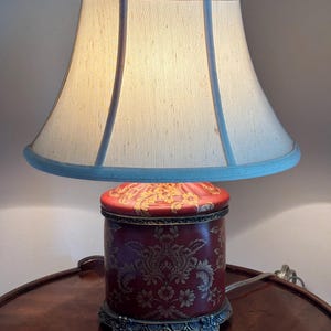 Może przedstawiać: Lampa stołowa z cylindryczną czerwoną podstawą w złoty wz&oacute;r damasceński, ozdobną metalową podstawą z dekoracyjnymi n&oacute;żkami oraz rozkloszowanym abażurem z tkaniny w kolorze złamanej bieli z jasnoniebieskim wykończeniem i małym zwieńczeniem w kształcie korony.
