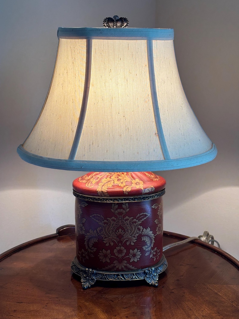 Może przedstawiać: Lampa stołowa z cylindryczną czerwoną podstawą w złoty wz&oacute;r damasceński, ozdobną metalową podstawą z dekoracyjnymi n&oacute;żkami oraz rozkloszowanym abażurem z tkaniny w kolorze złamanej bieli z jasnoniebieskim wykończeniem i małym zwieńczeniem w kształcie korony.